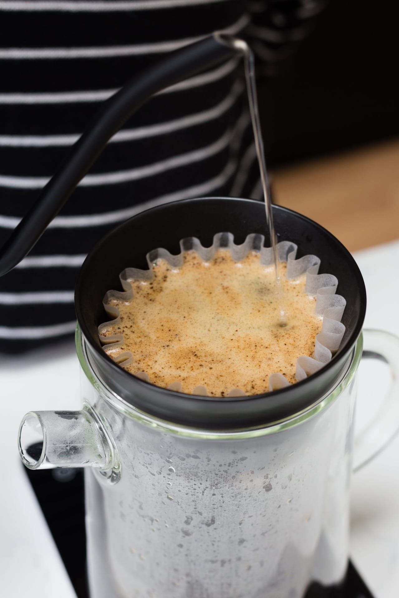 Close up of long kettle spout pouring boiling water into pour-over coffee mug with frothy coffee.