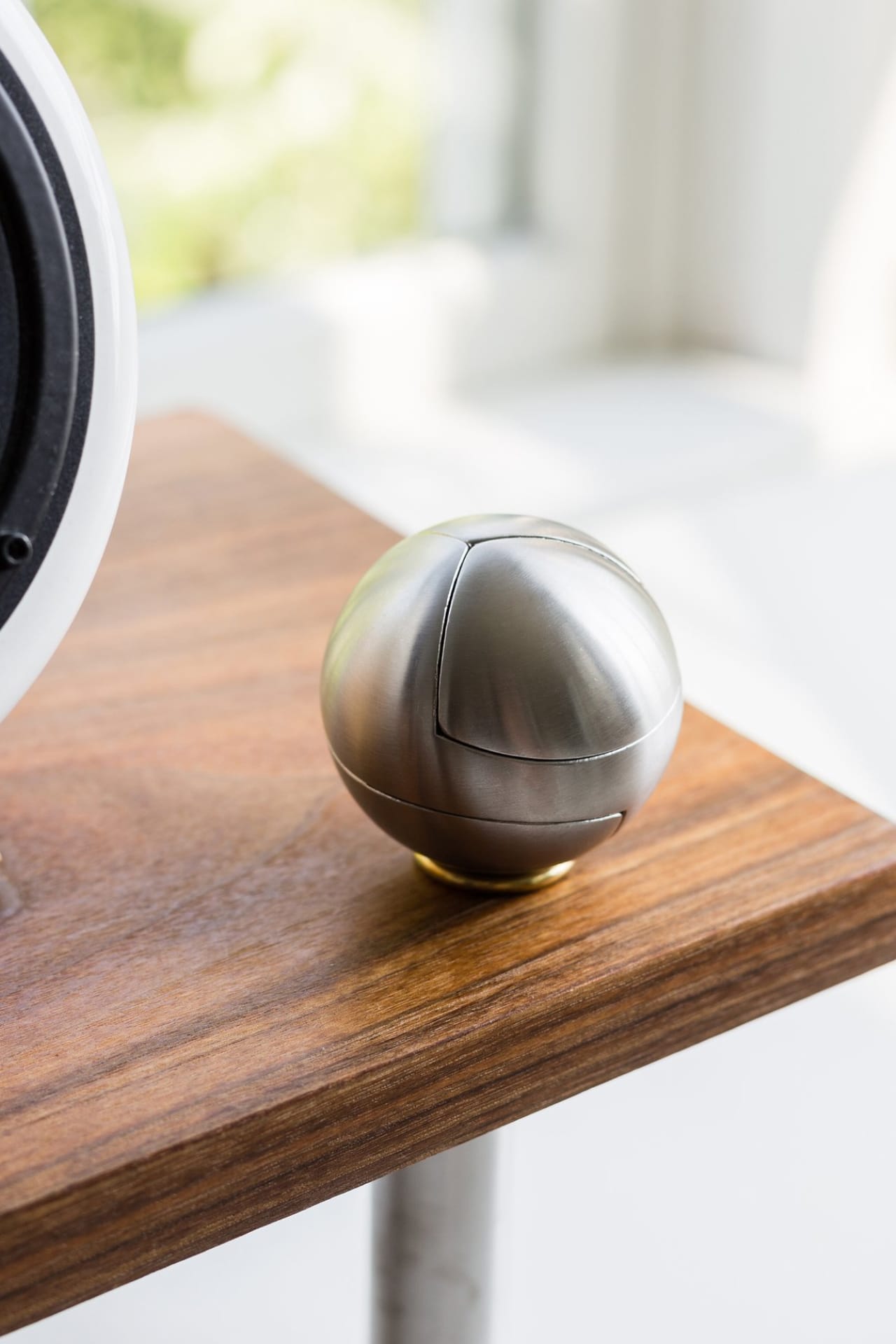 Machined steel sphere puzzle with smooth finish, geometric seams, and included brass stand on wood desk.