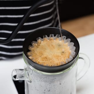 Kettle spout pouring boiling water into coffee grounds in pour-over mug.
