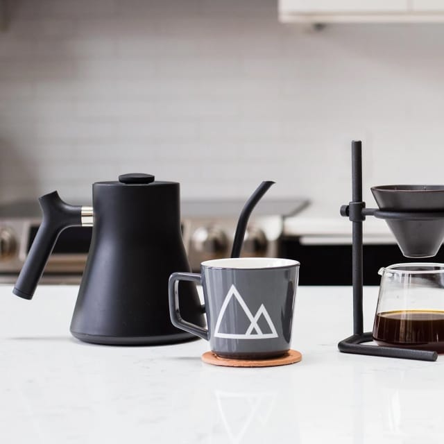 Black kettle with long pour spot and angled body on marble counter next to coffee mug and pour-over system.