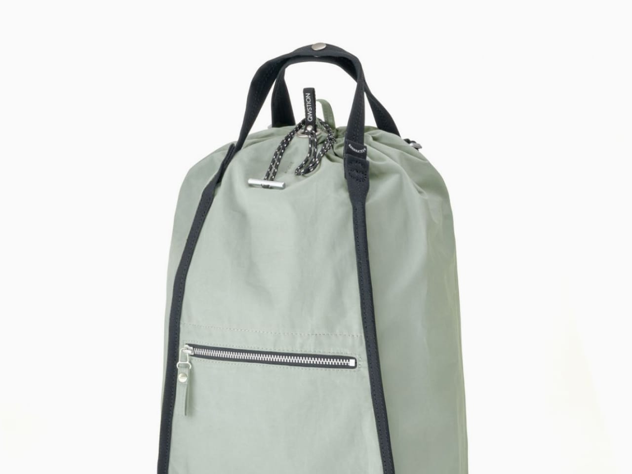 Light green canvas bag with black straps, handle, front zipper pouch, and drawstring top.